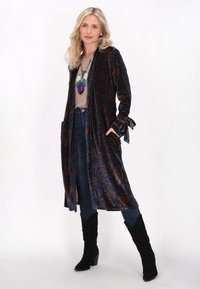 Lange, marineblauwe fluwelen kimono met een ornamentaal paisleypatroon, voorzien van zijzakken en striksluitingen bij de mouwen, gedragen over een beige top en jeans.