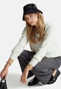 Ein weißer Strickpullover mit gerippten Bündchen, graue, maßgeschneiderte Hosen und ein schwarzer Bucket Hat. Schwarze Loafers und eine kleine schwarze Handtasche sind sichtbar.