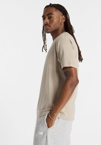 Tricou bej cu mânecă scurtă, din material moale, cu croi lejer. Asortat cu pantaloni de trening gri, prevăzuți cu buzunare laterale și talie elastică.