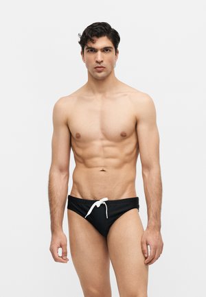 Sportos, sötét hajú férfi fekete úszónadrágot visel fehér zsinórral, egy sima világos háttér előtt áll, előre néz.