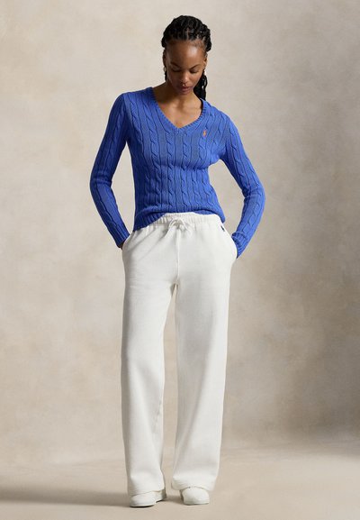Μπλε πλεκτό πουλόβερ με V-λαιμόκοψη και κεντημένο λογότυπο, συνδυασμένο με λευκή φαρδιά φόρμα και λευκά παπούτσια slip-on. Απαλά υφάσματα.