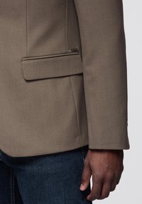 Brązowa marynarka w jodełkę z pojedynczą kieszenią i dopasowanym krojem. Tkanina ma gładką fakturę. Widoczne ręka spoczywająca na dżinsach.