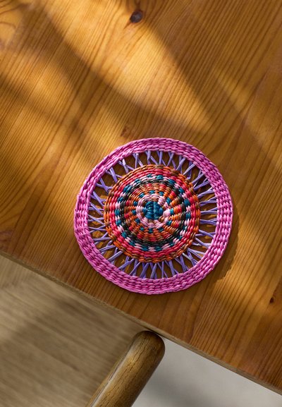 Round woven coaster in pink, purple, and multicolored fibers, featuring a central circular pattern and open weave detailing along the edges.