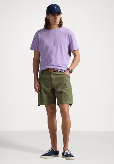 Lila bomullströja med broderad logotyp, olivgröna cargo-shorts, marinblå keps och blå sneakers med vita sulor. Avslappnad outfit.