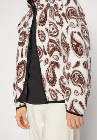 Fleecová bunda má bílou základnu s hnědými paisley vzory, zapínání na zip, elastické manžety a měkký, strukturovaný povrch.
