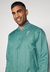Teal bomberjakke med en glat tekstur, lynlås lukning, sidelommer og broderet Liverpool FC logo i lys grå.