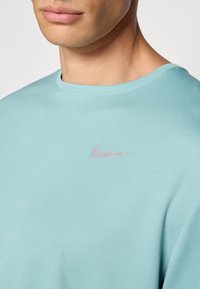 Lyseblå atletiktrøje med tekstureret stof, rund halsudskæring og et lille sølvfarvet Nike-logo på den øverste venstre side.
