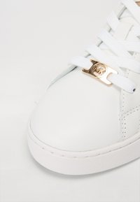 Balts ādas apavi ar gludu tekstūru, kas aprīkoti ar baltām auklām un zelta tonī metāla logotipa detalizāciju. Apaļā purngala un gumijas zoles projekts.