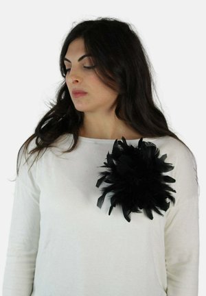 Femme aux longs cheveux noirs portant un haut blanc orné d'une grande broche noire à plumes sur la poitrine, regardant vers la gauche.