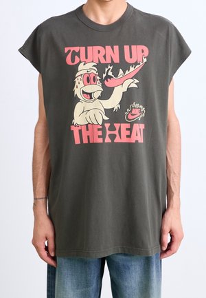 Homme portant un t-shirt sans manches gris foncé avec un texte rose « TURN UP THE HEAT » et un personnage de dessin animé tenant une chaussure Nike en flammes.