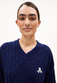 Dunkelblauer Strickpullover mit einem strukturierten Muster, V-Ausschnitt und weißem Logo-Akzent auf der linken Brust. Weicher Stoff mit gerippten Kanten.