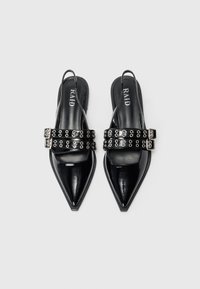 Mustat, kärjet ylöspäin suuntautuvat slingback-kengät, joissa on kaksoisvirityshihnat, joissa on hopeiset reiät, yksinkertaisella vaalealla taustalla.