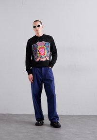 Svart tröja med färggrafisk design, marinblå byxor och svarta skor. Modellen bär vita solglasögon och står mot en ljusgrå bakgrund.