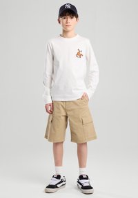 Chemise blanche à manches longues avec un graphisme orange, shorts cargo beiges et baskets noires avec des accents blancs. Casquette de baseball noire.