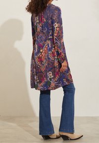 Tunika s květinovým vzorem v purpurové barvě, s dlouhými rukávy a A-silhouette, spárovaná s rozšířenými džíny a béžovými kotníkovými botami na podpatku.