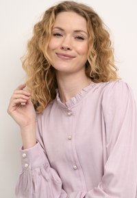 Levandulová blůza s volánovým límcem a knoflíkovým zapínáním. Dlouhé rukávy se nařasenými detaily. Hladká, lehká tkanina s perlovými akcenty.
