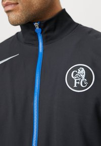 Crna sportska jakna s visokim ovratnikom, srebrni Nike logo, plavi patentni zatvarač i kružni CFC logo s grbom lava na prsima.