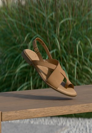 Sandaal van tan leer met gespband zwevend boven een houten bankje, groene gras wazig op de achtergrond.