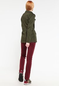 Osoba z jasnymi włosami w oliwkowej kurtce, bordowych spodniach i kamuflażowych trampkach idąca w stronę białego tła.