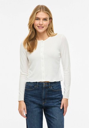 Jonge vrouw met lang blond haar, gekleed in een wit, met knopen gesloten, langärmelig shirt en blauwe spijkerbroek, staand tegen een effen achtergrond.