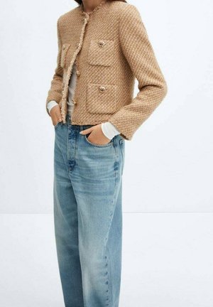 Personne portant une veste boutonnée texturée beige avec des poches sur une chemise blanche et un jean bleu clair ample, les mains dans les poches.