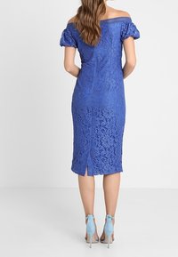 Modré krajkové šaty s odhalenými rameny a balonovými rukávy, délka ke kolenům, s květinovým vzorem a rozparkem na zádech; doplněné světle modrými podpatky.