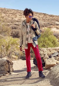 Beige buitenjack over een geruite shirt, rode broek en grijze wandelschoenen. Accessoires zijn een zonnebril en een blauwe rugzak. Woestijnlandschap op de achtergrond.