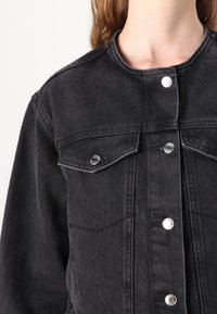 Črna denim jakna z ovratnikom, sprednjimi žepi, srebrnimi pritiskači in strukturiranim dizajnom z vidnimi šivi.