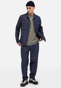 Marineblaue Jacke und Hose aus einem Stoff mit Blumenmotiven, kombiniert mit einem hellgrünen Strickpullover und schwarzen Schuhen.