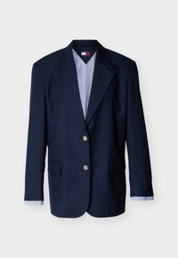 Μονόπατο σακάκι σε ναυτικό μπλε με δύο ασημένια κουμπιά, γιακά notch, τσέπες με καπάκι και ριγωτά μανίκια που φαίνονται στις μανσέτες του πουκαμίσου.