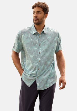 Hombre con cabello rizado y corto, que lleva una camisa de manga corta con botones y estampado verde claro, y pantalones oscuros, de pie con una mano en el bolsillo.