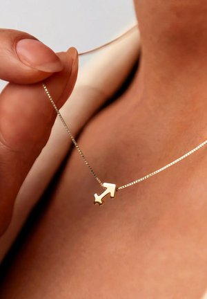 Hand holding a delicate gold chain necklace with a small Sagittarius zodiac arrow pendant resting on bare skin.