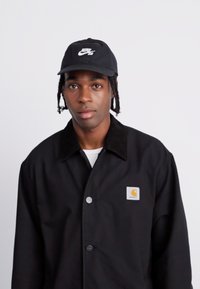 Crna jakna s oštrim ovratnikom i zatvaranjem na srebrni gumb, s kvadratnom oznakom. Crna kapa s bijelim logotipom.