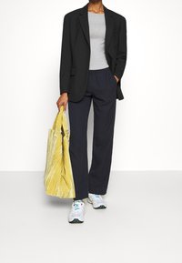 Černý blazer, šedé přiléhavé tričko, tmavě modré kalhoty a bílé tenisky. Třásňová taška žluté a bílé barvy s pruhy. Moderní, casual outfit pro různé příležitosti.