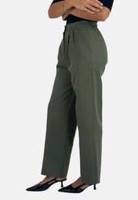Pantaloni verde oliva a vita alta con pieghe, tasche laterali e texture liscia, abbinati a tacchi a punta neri.