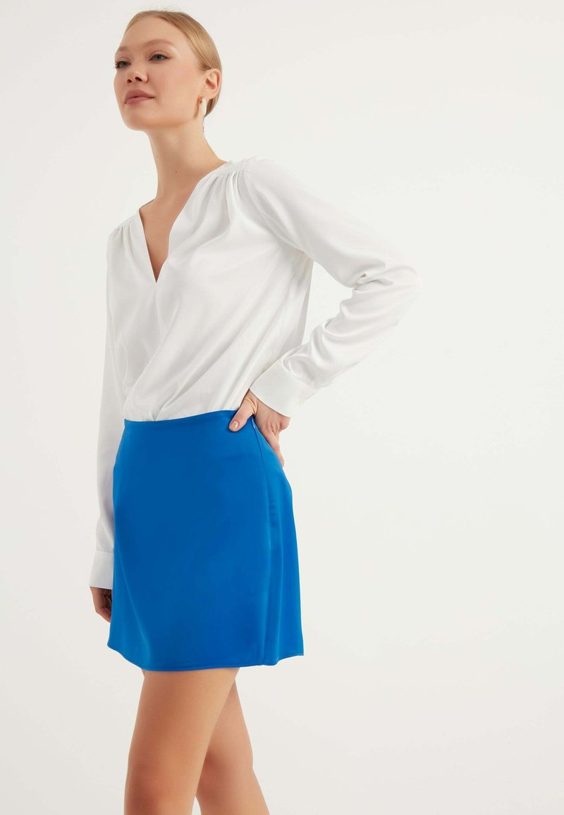 Vit långärmad blus med V-ringning, tillsammans med en klarblå minikjol. Blusen har en mjuk textur; kjolen är slät och lätt glänsande.