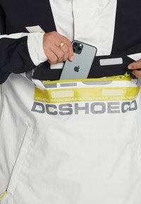 Hvid og sort jakke med en frontlomme med lynlås, der har et logo og en lys gul detalje. En telefon opbevares i lommen.