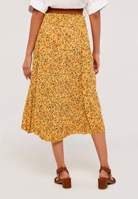 Blumenmaxirock in Gelb mit roten und grünen Mustern, mit gerafftem Taillenbund und fließendem Design. Kombiniert mit braunen Blockabsatzsandalen.