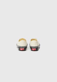 Canvas slip-on shoes in off-white with black sole and stripe. Features red logo on heel and minimal stitching details. Smooth texture.