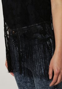 Svart spetsblus med fransdetaljer längs fållen, tillsammans med mörka, texturerade jeans. Spetsen har ett blommönster och en slät textur.