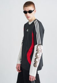 Μαύρο και κόκκινο αθλητικό μπλουζάκι με λευκά μανίκια που διαθέτει έντονη επιγραφή "adidas", ριγέ ώμους και ομαλή υφή υφάσματος. Συμπεριλαμβάνονται μαύρα γυαλιά ηλίου.