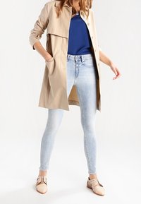 Abrigo trench beige con cuello muesca, dos bolsillos, combinado con jeans ajustados azul claro y zapatos beige con detalles a rayas.