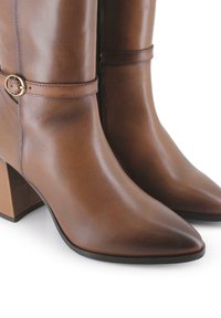 Botas de tobillo de cuero marrón con punta afilada, tacón cuadrado y una correa con una hebilla circular dorada. Textura suave, detalles mínimos.