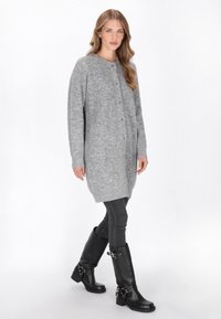 Femeie care merge îmbrăcată cu un cardigan lung gri cu nasturi, blugi skinny negri și cizme negre până la genunchi cu catarame, pe un fundal alb.