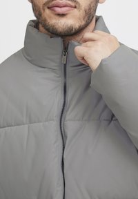 Gray padded jacket with a high collar and zipper front. Material appears smooth, with visible stitching and a relaxed fit around the shoulders.