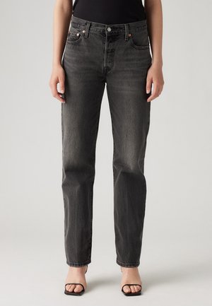 Personne portant un jean noir délavé taille haute, coupe droite, avec poches et braguette à boutons, associé à des sandales noires à talons et à brides.