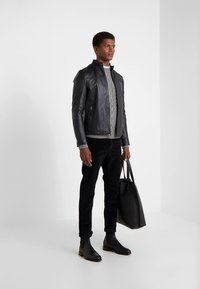 Black leather jacket, grey cable-knit sweater, black trousers, and black ankle boots. Model holds a black tote bag. Simple background.