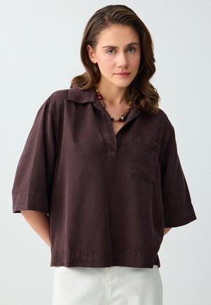 Femme aux cheveux bruns ondulés porte une chemise marron foncé oversize avec poche et un collier de perles colorées, debout devant un fond uni.