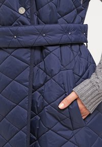 Marinblått kviltat väst med bälte i midjan, med tryckknappar och diamantformad söm; grå ribbad ärm synlig bredvid.