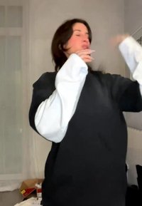 Giovane con lunghi capelli scuri che indossa maniche stratificate nere e bianche, gesticola all'interno di una stanza con una finestra e disordine.